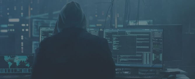 A man in a black hoodie working on two computers.
