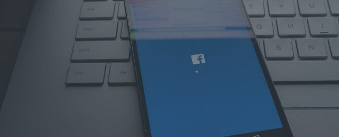 A phone with Facebook on the screen laying on top of a keyboard.