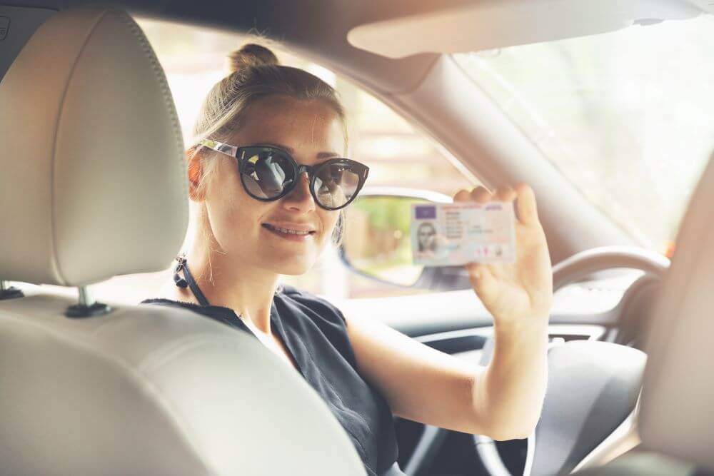 A girl sitting inside her car holding a driver's license.