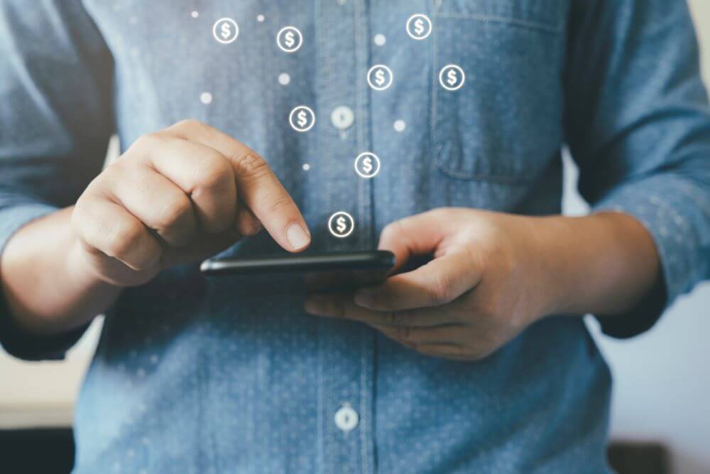 A man standing holding his smart phone with floating money icons.