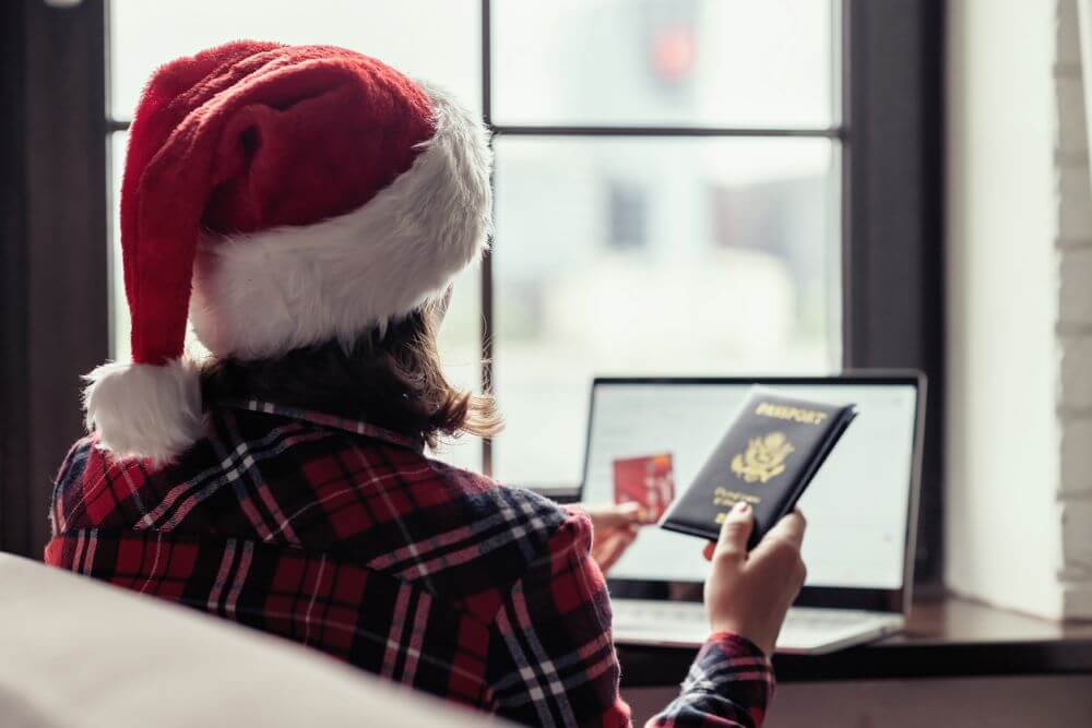 Woman in a red santa claus hat holding credit card and american passport using laptop for making order sitting near window.