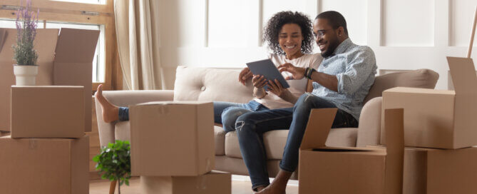 Couple sitting in house with moving boxes on tablet smiling.