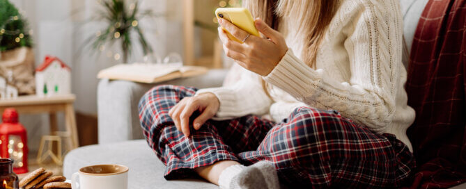 Cozy woman in knitted winter warm socks, sweater and checkered plaid with phone, drinking hot cocoa or coffee in mug resting on couch at home.