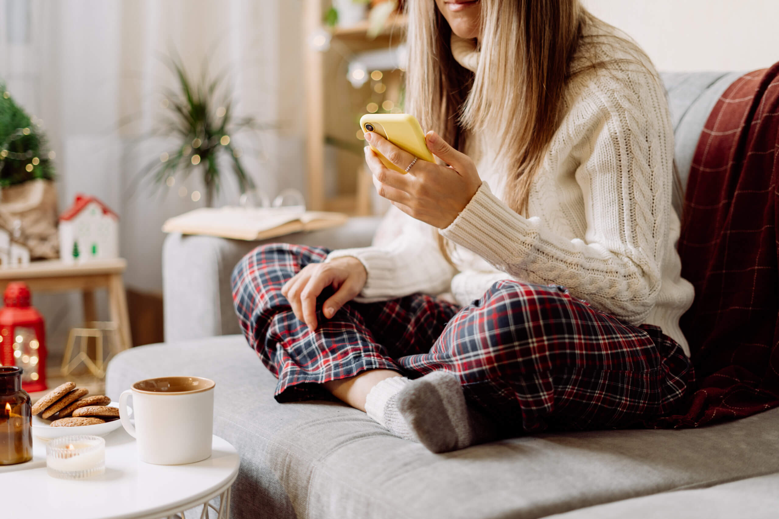 Cozy woman in knitted winter warm socks, sweater and checkered plaid with phone, drinking hot cocoa or coffee in mug resting on couch at home.